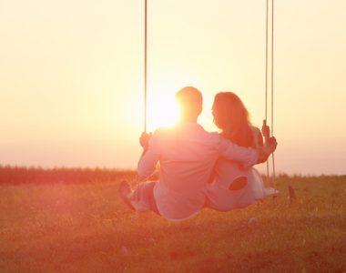 LENS FLARE: Happy couple swaying on a swing embraced on sunlit summer evening.